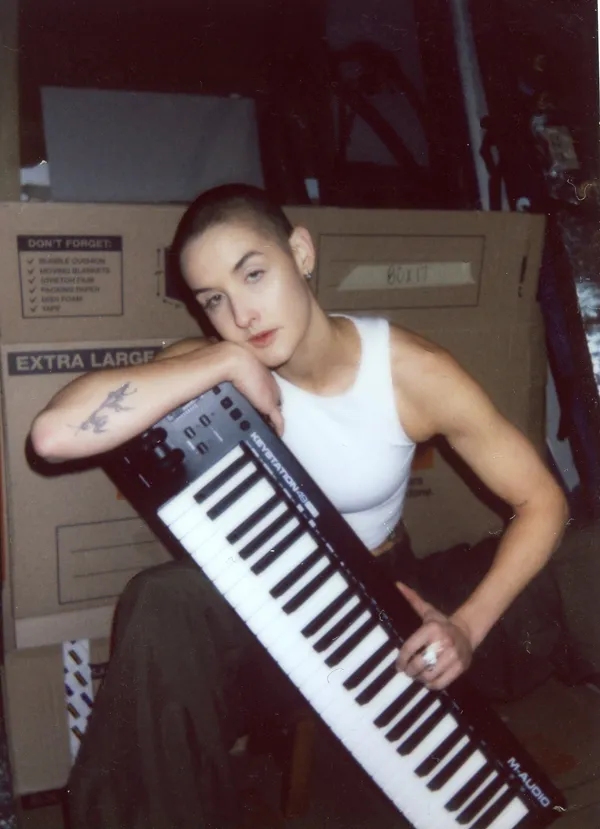 Madeline holds a digital keyboard vertically at an angle and rests their cheek against it, wearing a white beater tank with moving boxes in the background